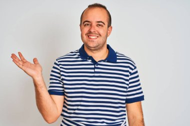 Sıradan çizgili polo giyen genç adam izole edilmiş beyaz arka planda dikiliyor neşeli bir şekilde gülümsüyor ve avuç içiyle kameraya bakıyor..