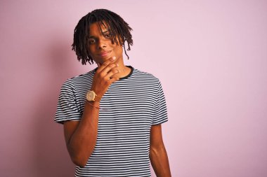 Rasta saçlı, donanma çizgili tişörtlü bir Afro adam izole edilmiş pembe arka planda dikiliyor kameraya bakıyor kolları çaprazlanmış ve elleri çenesinde kaldırılmış bir şekilde gülümsüyor. Olumlu düşünüyorum..