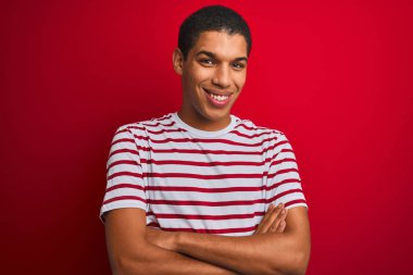 İzole edilmiş kırmızı arka plan üzerinde çizgili tişört giyen yakışıklı Arap adam kameraya bakarken kollarını kavuşturup gülümsüyor. Pozitif kişi.