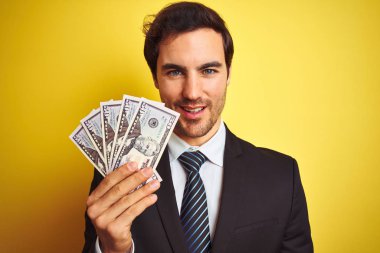 Genç yakışıklı işadamı ayakta mutlu bir yüz ile izole sarı arka plan üzerinde dolar tutan takım elbise giyen ve dişleri gösteren kendinden emin bir gülümseme ile gülümseyerek
