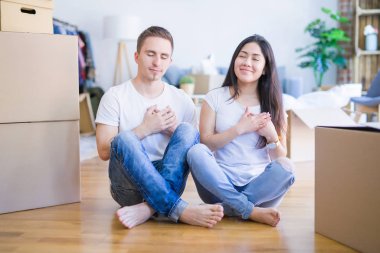 Young beautiful couple sitting on the floor at new home around cardboard boxes smiling with hands on chest with closed eyes and grateful gesture on face. Health concept.