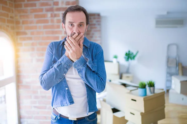 Middle age senior man moving to a new house packing cardboard boxes shocked covering mouth with hands for mistake. Secret concept.