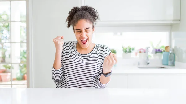 Beautiful african american woman with afro hair wearing casual striped sweater very happy and excited doing winner gesture with arms raised, smiling and screaming for success. Celebration concept.