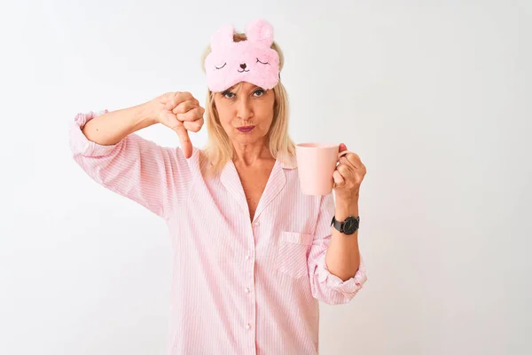 Middle age woman wearing sleep mask pajama drinking coffee over isolated white background with angry face, negative sign showing dislike with thumbs down, rejection concept