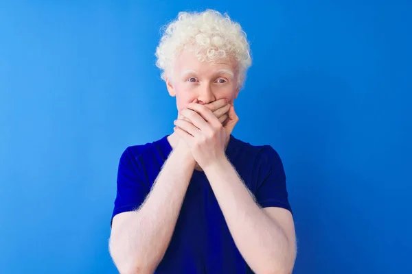 Young albino blond man wearing casual t-shirt standing over isolated blue background shocked covering mouth with hands for mistake. Secret concept.