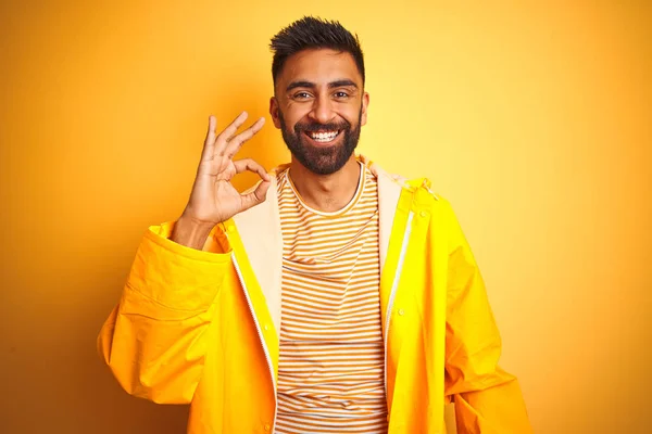 Young indian man wearing raincoat standing over isolated yellow background smiling positive doing ok sign with hand and fingers. Successful expression.