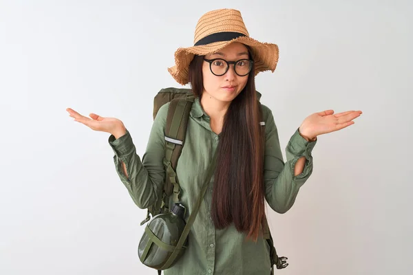 Chinese hiker woman wearing canteen hat glasses backpack over isolated white background clueless and confused expression with arms and hands raised. Doubt concept.