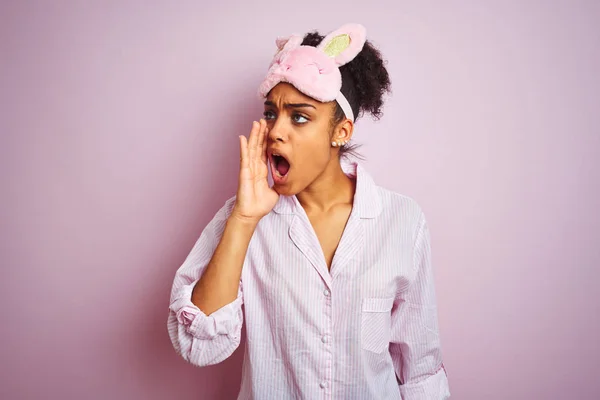 Young african american woman wearing pajama and mask over isolated pink background shouting and screaming loud to side with hand on mouth. Communication concept.