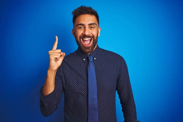 Young indian businessman wearing elegant shirt and tie standing over isolated blue background pointing finger up with successful idea. Exited and happy. Number one.