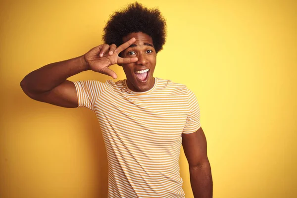 American man with afro hair wearing striped t-shirt standing over isolated yellow background Doing peace symbol with fingers over face, smiling cheerful showing victory