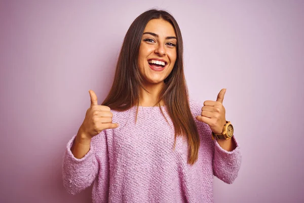 孤立したピンクの背景の成功の印の上に立つカジュアルセーターを身に着けている若い美しい女性は手で肯定的なジェスチャーをする 親指を笑顔と幸せ 陽気な表情と勝者のジェスチャー — ストック写真