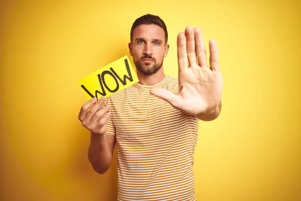 Young man holding wow text banner over yellow isolated background with open hand doing stop sign with serious and confident expression, defense gesture