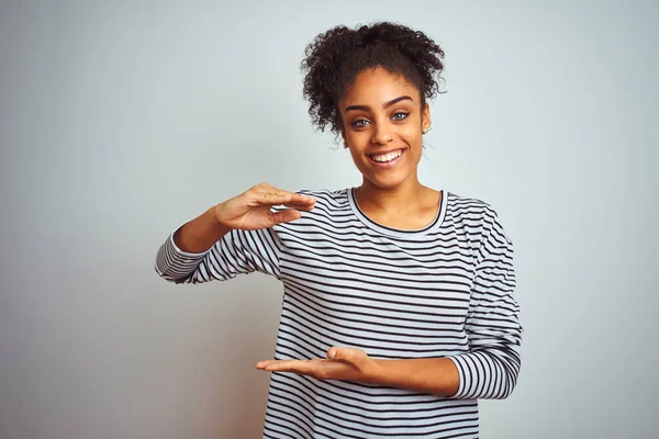 African american woman wearing navy striped t-shirt standing over isolated white background gesturing with hands showing big and large size sign, measure symbol. Smiling looking at the camera. Measuring concept.