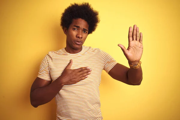 American man with afro hair wearing striped t-shirt standing over isolated yellow background Swearing with hand on chest and open palm, making a loyalty promise oath