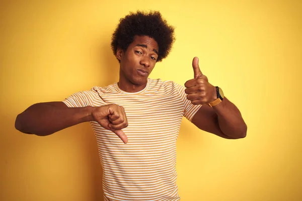 American man with afro hair wearing striped t-shirt standing over isolated yellow background Doing thumbs up and down, disagreement and agreement expression. Crazy conflict