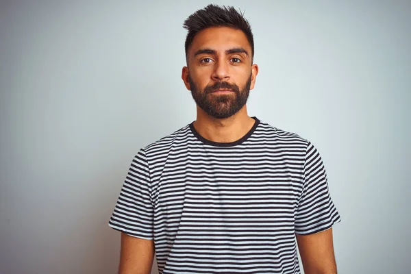 Young indian man wearing black striped t-shirt standing over isolated white background with serious expression on face. Simple and natural looking at the camera.