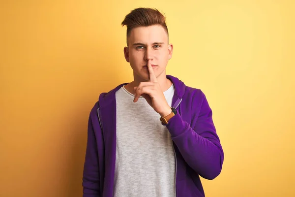 孤立した黄色の背景の上に立つ紫色のスウェットシャツを着た若いハンサムな男が唇に指で静かにするように求めている 沈黙と秘密の概念 — ストック写真