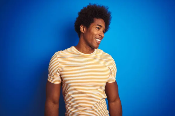 American man with afro hair wearing striped yellow t-shirt over isolated blue background looking away to side with smile on face, natural expression. Laughing confident.