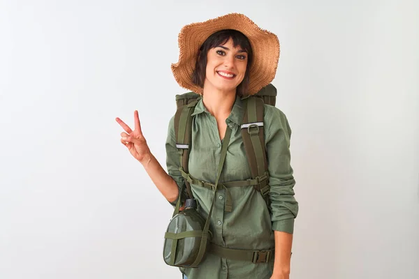 Hiker woman wearing backpack hat and water canteen over isolated white background smiling with happy face winking at the camera doing victory sign. Number two.