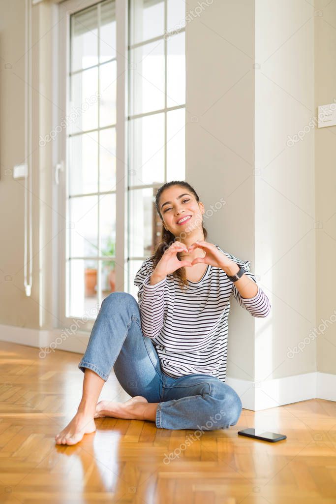 Young beautiful woman sitting on the floor at home smiling in love showing heart symbol and shape with hands. Romantic concept.