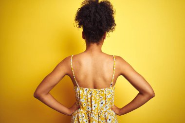 Siyah çiçekli bir elbise giyen Afro-Amerikalı bir kadın izole edilmiş sarı arka planda dikiliyor geriye bakıyor kolları vücudunda.