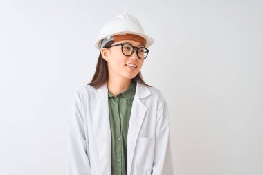 Young chinese engineer woman wearing coat helmet glasses over isolated white background looking away to side with smile on face, natural expression. Laughing confident.