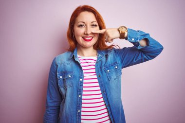 Güzel kızıl saçlı kadın kot gömlek ve çizgili tişört giyerek izole edilmiş pembe arka plan üzerinde el ve burun parmaklarıyla işaret ederek neşeli bir şekilde gülümsüyor. Güzellik konsepti