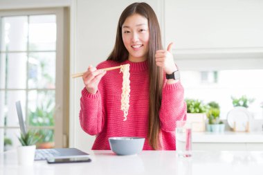 Güzel Asyalı kadın laptop kullanarak çalışan ve büyük bir gülümseme ile mutlu Asya noodle yeme ok işareti yapıyor, parmaklar ile başparmak, mükemmel işareti