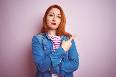 Güzel kızıl saçlı kadın kot gömlek ve çizgili t-shirt ile izole edilmiş pembe arka plan üzerinde el ile işaret ederek reklamı gösteriyor, ciddi ve sakin yüz.