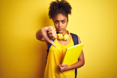 Sırt çantası ve kulaklık kullanan Afro bir kadın izole edilmiş sarı arka plan üzerinde not defteri tutuyor. Kızgın yüz ifadesi, olumsuz işaret baş parmakları aşağıda, reddedilme konsepti.