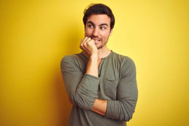 İzole edilmiş sarı arka plan üzerinde sıradan tişörtler giyen genç yakışıklı adam elleri ağız yiyen tırnaklarla gergin ve stresli görünüyor. Endişe sorunu.