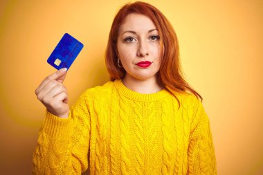 Genç müşteri kızıl saçlı kadın kredi kartını sarıya tutarak izole edilmiş bir arkaplan kullanıyor. Akıllı bir yüz ifadesiyle ciddi olduğunu düşünüyor.