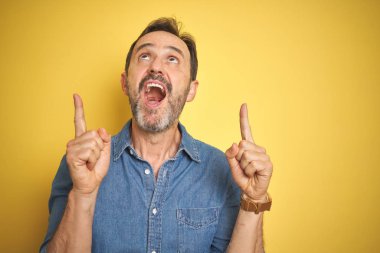 Sarı arka planda gri saçlı yakışıklı orta yaşlı bir adam şaşırmış ve şaşırmış bir şekilde parmaklarıyla işaret ediyor ve kollarını kaldırıyordu..