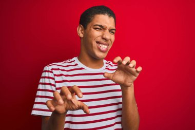 İzole edilmiş kırmızı arka plan üzerinde çizgili tişört giyen genç yakışıklı Arap adam kedi gibi komik pençe hareketleri yapıyor, agresif ve seksi bir ifade.