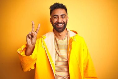 Young indian man wearing raincoat standing over isolated yellow background showing and pointing up with fingers number two while smiling confident and happy.