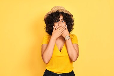 Young arab woman with curly hair wearing t-shirt and hat over isolated yellow background shocked covering mouth with hands for mistake. Secret concept.