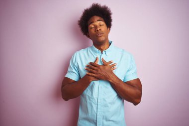 Afro saçlı, mavi gömlekli genç bir adam izole edilmiş pembe arka plan üzerinde duruyor elleri göğsünde, gözleri kapalı ve yüzünde minnet dolu bir jest ile gülümsüyor. Sağlık kavramı.
