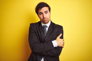 Takım elbiseli ve kravatlı yakışıklı genç bir işadamı izole edilmiş sarı arka planda duruyor. Kış soğuğu için titriyor ve donuyor. Yüzünde üzgün ve şok ifadesi var.