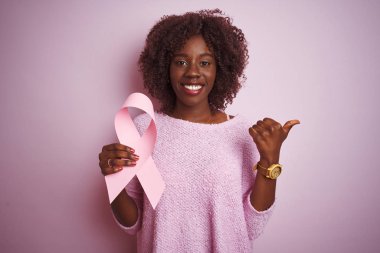 Genç Afrikalı Afro kadın kanserli kurdele tutuyor pembe arka planda duruyor ve gülümseyen bir gülümsemeyle baş parmağıyla işaret ediyor.