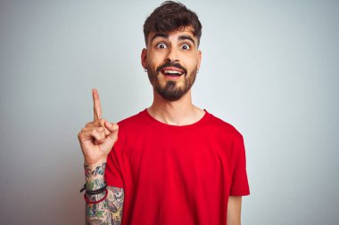 İzole edilmiş beyaz arka planda duran kırmızı tişörtlü genç adam başarılı bir fikirle işaret ediyor. Heyecanlı ve mutlu. Bir numara..