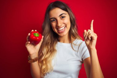 Young beautiful woman holding pepper over red isolated background very happy pointing with hand and finger to the side