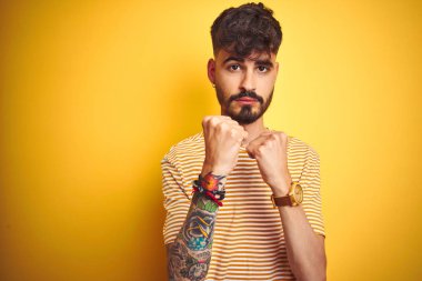 Dövmeli, çizgili tişörtlü genç adam izole edilmiş sarı arka planda duruyor. Yumruk savunma hareketleriyle dövüşmeye hazır, kızgın ve üzgün yüzlü, sorundan korkan.