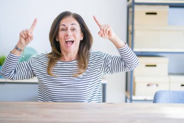 Orta yaşlı bir kadın evde masada oturuyor. Şaşırmış, şaşırmış, parmaklarıyla işaret ediyor ve kollarını kaldırıyor..