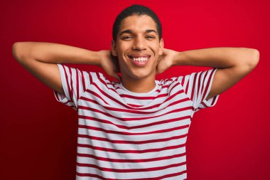Genç yakışıklı Arap adam izole edilmiş kırmızı arka planda çizgili tişört giyiyor rahatlıyor ve geriliyor, kolları ve elleri başının arkasında mutlu bir şekilde gülümsüyor.
