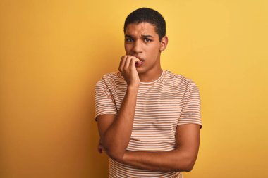 İzole edilmiş sarı arka plan üzerinde duran çizgili tişört giyen genç, yakışıklı Arap adam elleri ağız yiyen tırnaklarla stresli ve gergin görünüyor. Endişe sorunu.