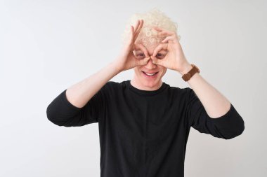 Siyah tişörtlü genç albino bir adam izole edilmiş beyaz arka planda dikiliyor. Dürbün gibi jest yapıyor, gözleri parmaklarından bakıyor. Çılgın ifade.