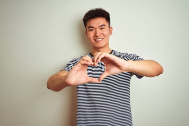 Asyalı, çizgili tişört giyen genç bir Çinli, izole edilmiş beyaz arka planda duruyor. Aşkla gülümsüyor, kalp sembolü ve şekli elleriyle gösteriyor. Romantik konsept.