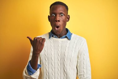 African american man wearing denim shirt and white sweater over isolated yellow background Surprised pointing with hand finger to the side, open mouth amazed expression.