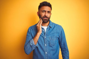 Young indian man wearing denim shirt standing over isolated yellow background touching mouth with hand with painful expression because of toothache or dental illness on teeth. Dentist concept.