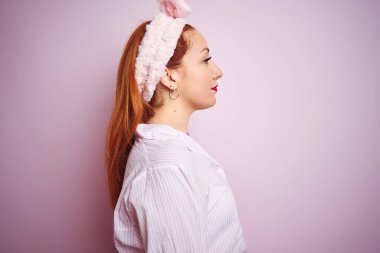 Genç, güzel, kızıl saçlı bir kadın pembe izole edilmiş arka planda dikiliyor. Yan yana bakıyor, rahat bir görünüm sergiliyor. Kendine güvenen bir gülüşü var..